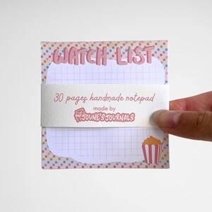 Peut inclure: Bloc-notes fait à la main avec un design rose et à pois. Le bloc-notes porte les mots "WATCH LIST" en haut et comprend 30 pages. Un dessin de pop-corn est en bas à droite. Fabriqué par JOUNE'S JOURNALS.