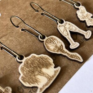 May include: Close-up of several wooden earrings with bronze-colored ear wires. The earrings feature various designs, including a tree, champagne glasses, and a floral shape. The earrings are light brown with dark brown edges.