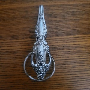 May include: A silver spoon handle with a decorative floral design, attached to a circular ring. The spoon handle is ornate with detailed patterns and curves. The ring is smooth and simple, contrasting with the handle's intricate design. The object is likely a unique key ring or decorative item.