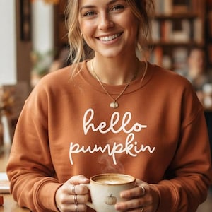 Peut inclure: Un sweat-shirt à manches longues de couleur rouille avec le texte blanc en cursive hello pumpkin sur le devant. Le vêtement présente une encolure ronde et est porté avec un délicat collier à chaîne dorée. Le tissu semble être un mélange de coton doux.