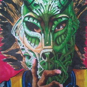 Könnte beinhalten: Eine grüne und goldene Maske mit komplizierten Details, die das Gesicht einer Person mit brauner Haut bedeckt. Die Maske hat ein scharfes, kantiges Design mit einer spitzen Oberseite und einer langen, schmalen Nase. Die Hand der Person ist im Vordergrund sichtbar und hält die Maske fest.