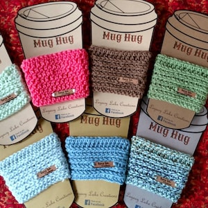 May include: Six crocheted mug huggers in various colors, including pink, brown, green, and blue. Each mug hugger has a small wooden tag with the text "Mug Hug" and "Logany Lake Creations".