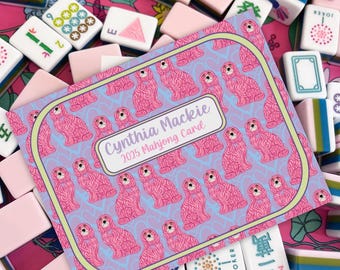 May include: A collection of pink and white Mahjong tiles and a Cynthia Mackie 2025 Mahjong Card. The card features a repeating pattern of pink poodle illustrations on a light blue background, framed with a green border.