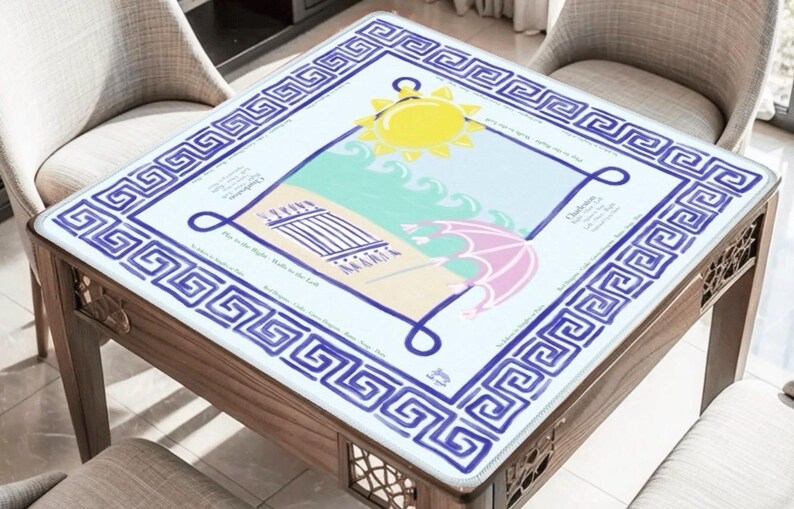 Puede incluir: Mesa cuadrada de madera con un dise&ntilde;o de escena de playa en la parte superior. El dise&ntilde;o incluye un sol amarillo, un paraguas rosa y olas azules. La mesa tiene un borde de clave griega azul y blanco.