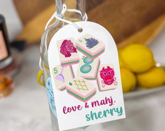 May include: A clear glass bottle with a white tag featuring colorful Mahjong tiles and the text "love & mahj. sherry." The tag is tied to the bottle with white string. The bottle has a label that says "JE Distillery."