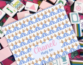Puede incluir: Un conjunto de fichas de Mahjong rosas y blancas con una tarjeta decorativa. La tarjeta presenta un patrón de paraguas azules y ardillas marrones, con el texto "Chanel Mahj On" en rosa.