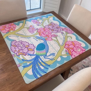 May include: A square, colorful placemat featuring a whimsical design. The placemat has a light blue background with a stylized bird, pink and purple flowers, and green branches. Text is included in the design. The placemat is on a wooden table.