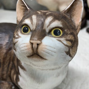 May include: A ceramic figurine of a brown and white tabby cat with yellow eyes. The cat is lying down with its head raised and looking directly at the viewer.