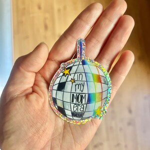 May include: A silver glitter disco ball sticker with a rainbow stripe and the text "In my mom era" in black lettering.