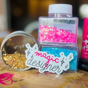 Könnte beinhalten: Zwei durchsichtige Plastikbehälter gefüllt mit pinkfarbenem und blauem Glitzer. Ein Aufkleber mit dem Text "magic designer" ist vor den Behältern platziert.