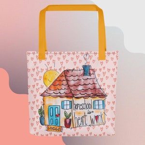 May include: A pink tote bag with a yellow handle and a cartoon illustration of a house with a red roof, a chimney, and a sun. The house has a blue door with a red heart and the text "homeschool is heart work." The background is a pink pattern of small red hearts.