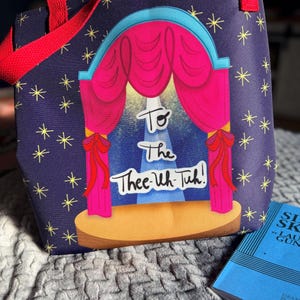 May include: A navy blue tote bag with a stage design featuring pink curtains, a blue arch, and the text "To The Thee-Uh-Tuh!". The bag has red handles and is decorated with yellow stars.