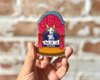 Peut inclure: Une broche en acrylique pailleté représentant une scène avec des rideaux roses, un sol jaune et un fond bleu avec un projecteur. Le texte "To The Thee-Uh-Tuh!" est imprimé en écriture blanche au centre, entouré d'une bordure scintillante.