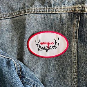 Puede incluir: Un parche ovalado blanco con un borde rosa cosido en una chaqueta vaquera azul. El parche tiene texto negro que dice "magic designer" con pequeños símbolos en forma de estrella a cada lado del texto.