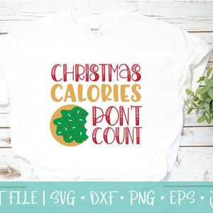Puede incluir: Camiseta blanca con texto rojo y dorado que dice "Las calorías de Navidad no cuentan". Una dona verde y blanca con chispas rojas está a la izquierda del texto.