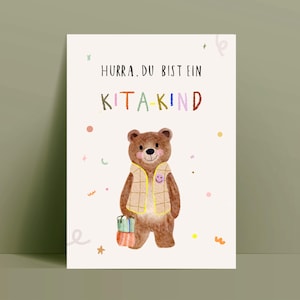 Könnte beinhalten: Aquarell-Illustration auf einer cremefarbenen Karte mit einem braunen Bären, der eine karierte Weste trägt und ein Geschenk hält. Der Text "HURRA. DU BIST EIN KITA-KIND" steht in Schwarz und bunten Buchstaben über dem Bären.