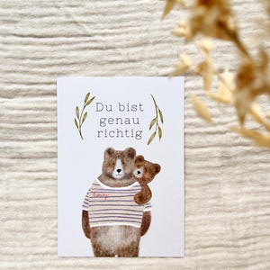 May include: A greeting card featuring a watercolor illustration of two bears. The larger bear wears a striped shirt and embraces a smaller bear. The German text "Du bist genau richtig" is at the top, with decorative leaves.