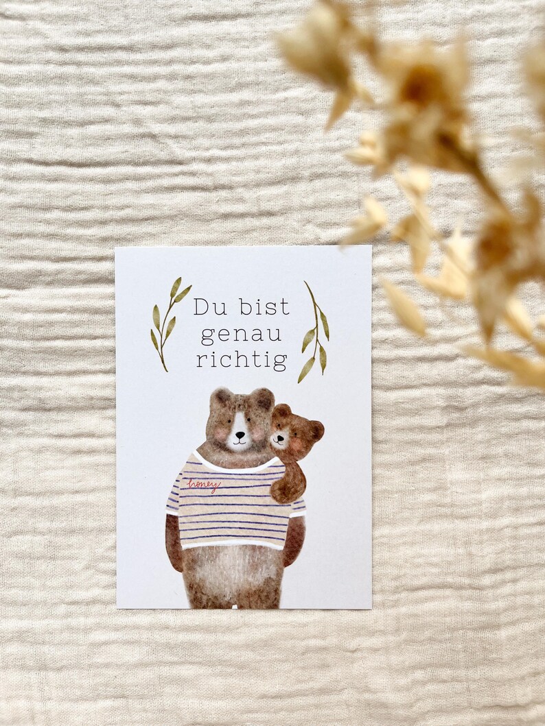 May include: A greeting card featuring a watercolor illustration of two bears. The larger bear wears a striped shirt and embraces a smaller bear. The German text "Du bist genau richtig" is at the top, with decorative leaves.