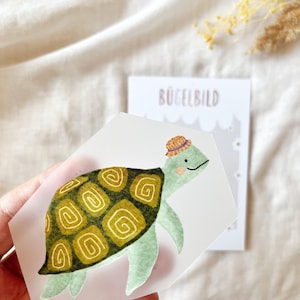 Könnte beinhalten: Eine Illustration einer Schildkröte mit Strohhut und grün-gelbem Panzer. Das Bild befindet sich auf einem transparenten Aufkleber, der von einer Hand gehalten wird. Im Hintergrund ist eine Karte mit dem Text "BÜGELBILD" zu sehen.