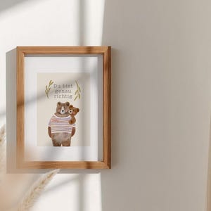 May include: A framed print featuring a watercolor illustration of two teddy bears. One bear wears a striped shirt and holds a smaller bear. The print includes the text "Du bist genau richtig" and is set in a wooden frame.