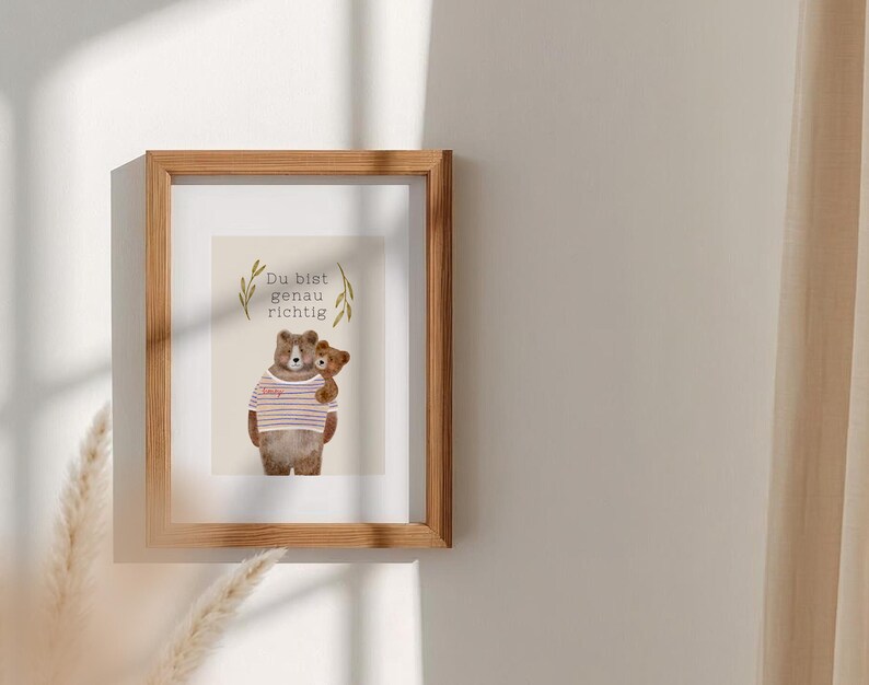 May include: A framed print featuring a watercolor illustration of two teddy bears. One bear wears a striped shirt and holds a smaller bear. The print includes the text "Du bist genau richtig" and is set in a wooden frame.