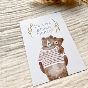 May include: A greeting card featuring a watercolor illustration of two brown bears. The larger bear wears a striped shirt with the word "honey" on it. The smaller bear is hugging the larger bear. The card has the text "Du bist genau richtig".