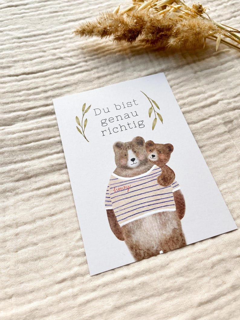 May include: A greeting card featuring a watercolor illustration of two brown bears. The larger bear wears a striped shirt with the word "honey" on it. The smaller bear is hugging the larger bear. The card has the text "Du bist genau richtig".