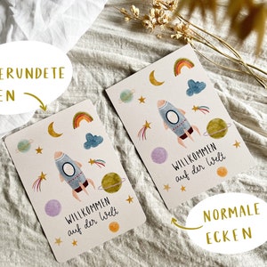 May include: Two greeting cards with a space theme. Each card features a rocket ship, planets, stars, rainbows, and clouds. The text "Willkommen auf der Welt" is printed on each card. One card has rounded corners, the other has normal corners.