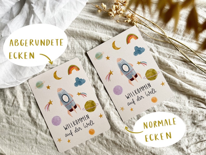 May include: Two greeting cards with a space theme. Each card features a rocket ship, planets, stars, rainbows, and clouds. The text "Willkommen auf der Welt" is printed on each card. One card has rounded corners, the other has normal corners.