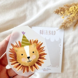 Könnte beinhalten: Aquarell-Illustration eines Löwengesichts mit Partyhut. Der Löwe hat eine braune Mähne, ein hellbraunes Gesicht und einen weißen Streifen. Das Bild befindet sich auf einem transparenten Aufkleber, mit einer Karte mit der Aufschrift "BÜGELBILD" im Hintergrund.