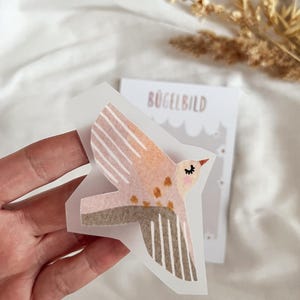 Könnte beinhalten: Ein rosa-brauner Bügelbild-Aufkleber mit einem Vogel mit geschlossenen Augen. Der Aufkleber befindet sich auf einer durchsichtigen Folie mit dem aufgedruckten Wort "Bügelbild".