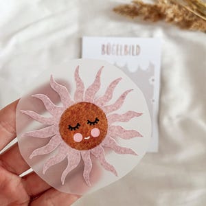 Könnte beinhalten: Eine rosa Filzsonne mit einem braunen Gesicht, geschlossenen Augen und rosa Wangen. Die Sonne befindet sich auf einer durchsichtigen Plastikfolie mit dem aufgedruckten Wort "Bügelbild".