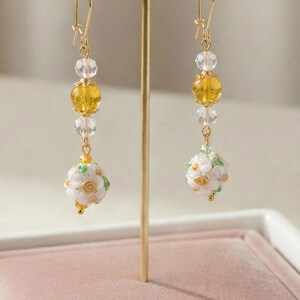 May include: A pair of dangling earrings with a gold-tone hook, featuring clear and yellow beads, and a white floral ball at the bottom. The earrings are displayed on a gold stand with a pink velvet base. The word "Glimmer" is on a small white card.