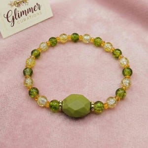 May include: A beaded bracelet with olive green and yellow beads, and a large olive green faceted center bead. The bracelet is on a pink surface, with a small card that reads "Glimmer Curations".