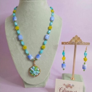 May include: A necklace and earring set featuring light blue rectangular beads, yellow star-shaped beads, and small green accent beads. The necklace includes a round gold-toned pendant filled with green and blue beads. A card reads Glimmer Curations.