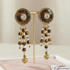 May include: A pair of dangle earrings with a circular, brown and gold design. Each earring features a central clear crystal, surrounded by a patterned brown border. Below, strands of brown beads and clear crystals cascade to a decorative gold-colored ball.