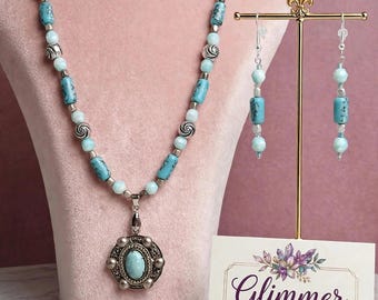 May include: A necklace and earring set featuring light blue and turquoise-colored cylindrical and round beads with silver-toned accents. The necklace includes a central oval pendant. A card displays the text Glimmer Curations.