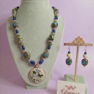 May include: A necklace and earring set featuring blue and green ceramic beads with silver-tone accents. The necklace includes a round pendant with a painted bird and floral design. The earrings feature matching beads and a small blue bead on a gold-tone stand.