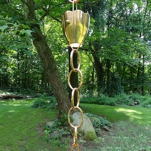 May include: A gold rain chain with two copper cups hanging from it. The chain is made of interlocking rings and the cups are shaped like bells. The chain is hanging from a tree branch in a forest setting.
