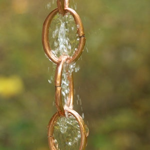 May include: A copper rain chain with water flowing through the links. The chain is made of multiple connected rings.