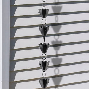 May include: A black metal rain chain with a series of flower-shaped cups hanging from a chain. The chain is attached to a white house with gray siding.