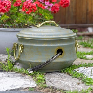 May include: A green and gold metal hose reel with a decorative lid and brass handles. The hose is black and is partially wound around the reel.