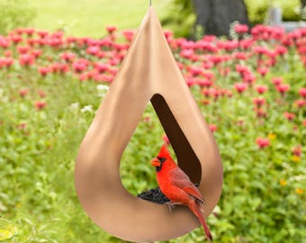 May include: A copper-colored teardrop-shaped bird feeder with a cardinal perched on the edge. The feeder is hanging from a wire and is surrounded by green foliage.