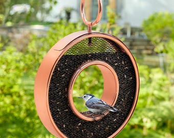 May include: A copper-colored bird feeder shaped like a ring with a smaller ring in the center. The feeder is filled with birdseed and a small bird is perched on the inner ring.