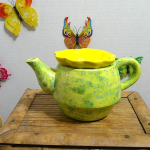 Peut inclure: Une théière en céramique avec un couvercle jaune, peinte dans un motif vert et bleu moucheté. La théière est posée sur une surface en bois, avec des décorations de papillons colorés sur le mur derrière elle.