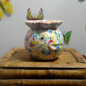 May include: A round ceramic bowl with a wide, scalloped rim, featuring a speckled, multi-colored glaze in shades of blue, yellow, pink, and red. The bowl has a smooth, rounded body with a small, raised, beak-like protrusion on one side.