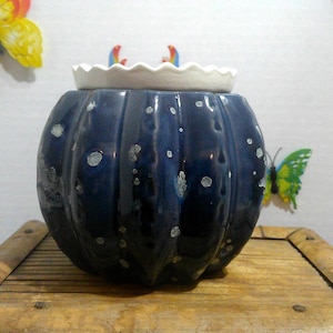 May include: A round, dark blue ceramic jar with a ribbed, pumpkin-like texture and a glossy finish featuring small, light-colored speckles. The jar is topped with a white, scalloped-edge lid that sits flush against the top opening of the vessel.