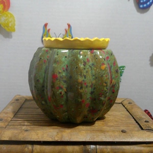 May include: A round, pumpkin-shaped ceramic jar with a glossy green glaze featuring multi-colored speckles in red, yellow, and orange. The jar is topped with a bright yellow, scalloped-edge lid that fits securely into the top opening of the vessel.