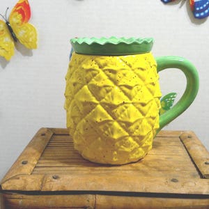 May include: A yellow pineapple-shaped ceramic mug with a green handle and rim, sitting on a wooden surface. Decorative butterfly accents are on the wall behind the mug. The mug is approximately 5 inches tall.