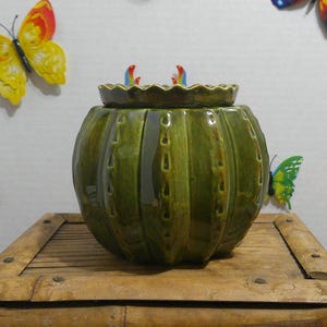 May include: A round, olive green ceramic jar with a ribbed, cactus-like texture and a scalloped lid. The jar features vertical ridges with small, indented details along the sides, finished with a glossy glaze that reflects light across its surface.
